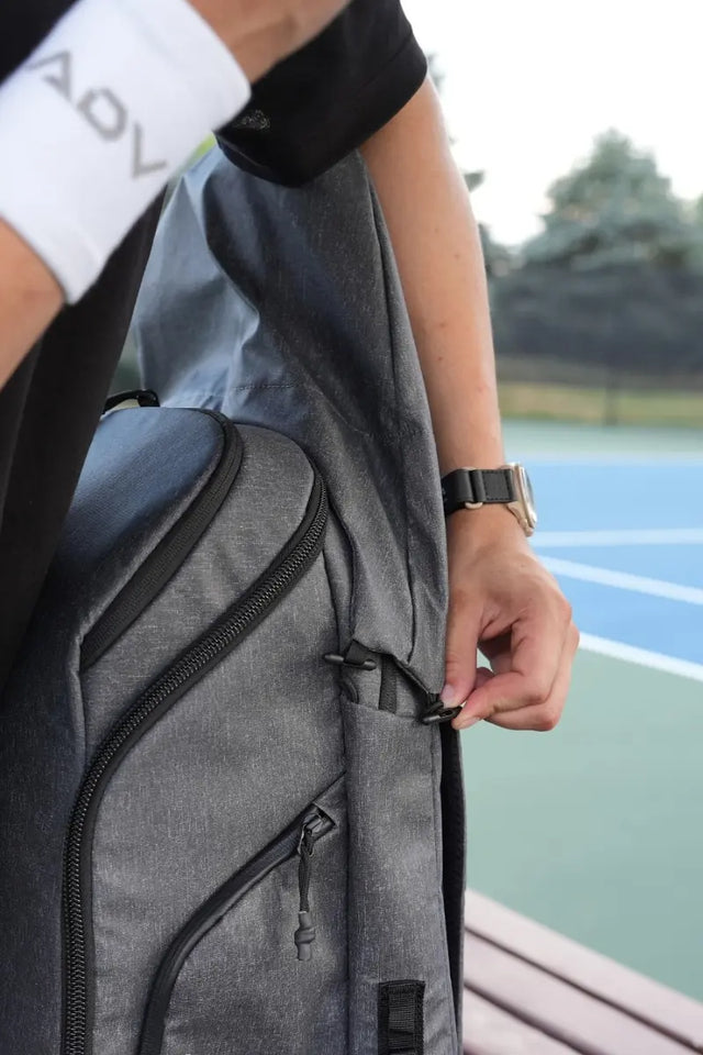 Sports gear for tennis includes a gray tennis backpack with a racket compartment, worn by a player near a court