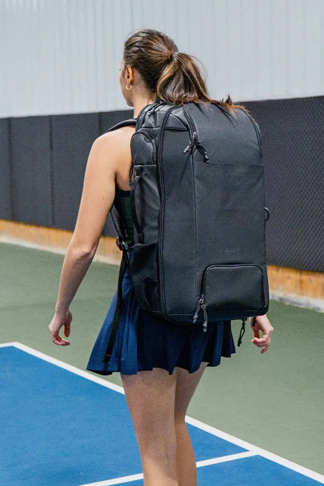 Competitive tennis gear including a black backpack worn by a female player on an indoor court