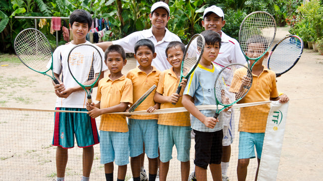 Smiling kids with tennis rackets lined up near a net with a pro tennis bag to the side.