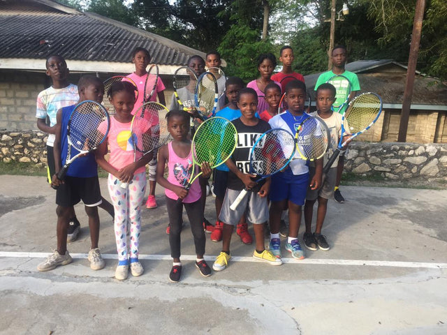 Group of children holding ADV tennis rackets on a tennis court, posing for a team photo outdoors.