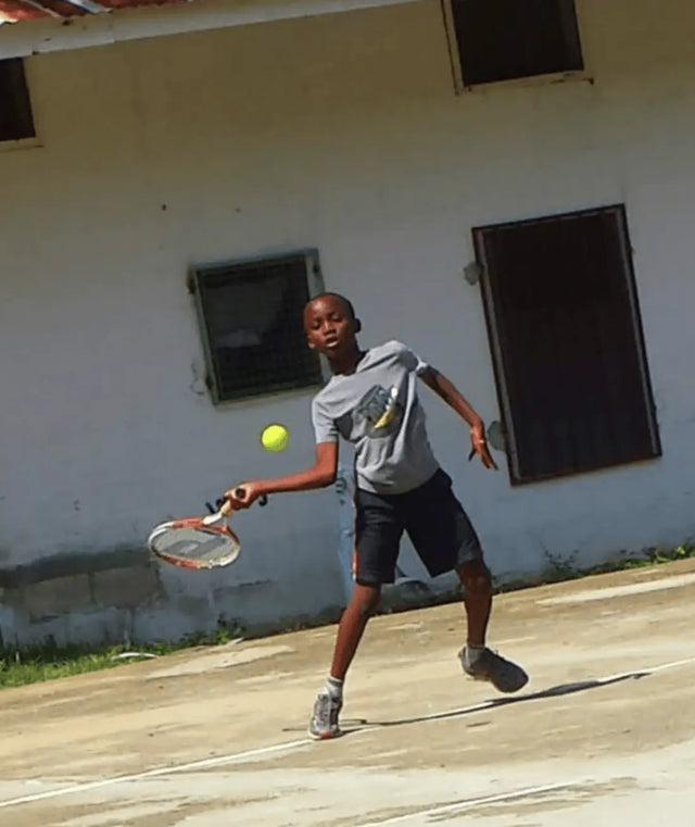 Boy swinging a tennis racket mid-match with a tennis racket backpack in the background near a wall.