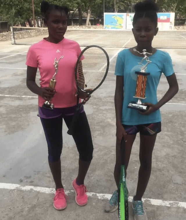 Tournament tennis bag sits behind two young girls holding trophies and tennis rackets on an outdoor court.