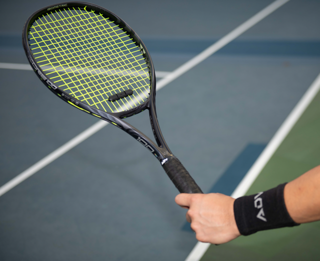 White male holding a tennis racket that has tennis overgrip on it. Also man is wearing tennis wristband