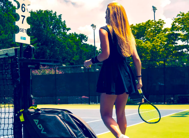 blonde female tennis player with a black tennis wristband standing on a tennis court holding racket with with white tennis overgrip. Next to the tennis net is placed tennis bag