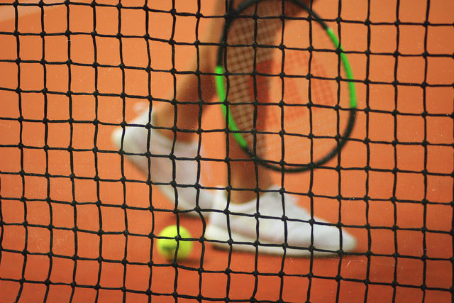 A person standing behind a tennis net holding a racket in his right hand. Green tennis ball sitting down on a tennis court