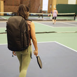 Pickleball bag worn by a player walking onto an indoor court, featuring a sleek black design with spacious compartments.